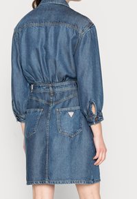 Denim-mekko kesk blue-värissä, jossa on puffihihat, vyötäröllä vyö ja kaksi takataskua kolmion muotoisella logopainatuksella.