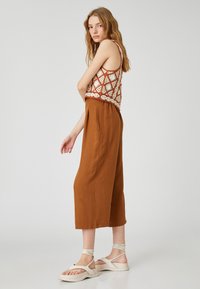 Brązowe spodnie w krótkim kroju o luźnym fasonie, zestawione z jasnym topem na ramiączkach w wzory w kolorze kremowym i rdzawej czerwieni. Model nosi jasne sandały z zapięciem na kostce.