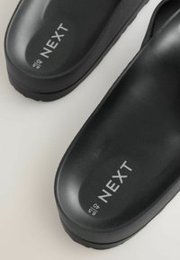 Schwarze Slip-on Sandalen mit dicken Sohlen, versehen mit der Aufschrift „NEXT“ und Größe 6,5/40, auf hellem grauem Hintergrund dargestellt.