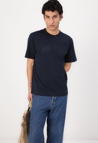 Μπλέ κοντομάνικο βαμβακερό t-shirt σε ναυτικό μπλε χρώμα με το κείμενο "BELSTAFF" σε ελαφρώς ανάγλυφη, τονική εκτύπωση. Συνδυάζεται με μπλε τζιν παντελόνι.