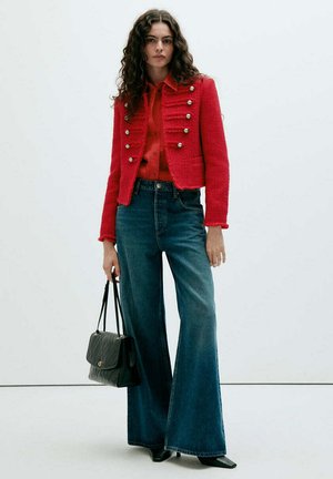 Mujer con cabello rizado que lleva una chaqueta roja abotonada, camisa roja, vaqueros anchos azules, zapatos negros y sostiene un bolso negro.