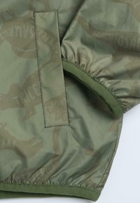 Grønt stof med et dinosaurmønster og en glat tekstur. Har elastiske manchetter, en lap og en fleksibel kantdetalje.