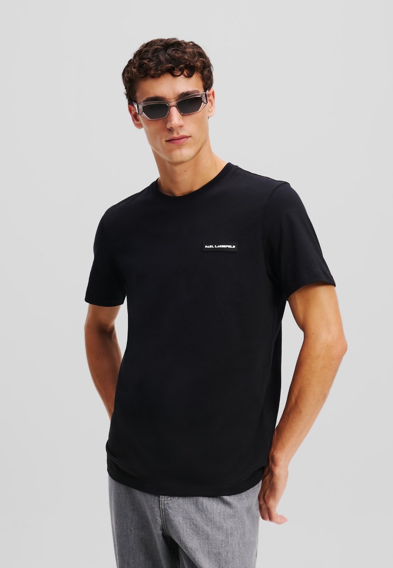 Tricou negru cu guler rotund, confecționat din bumbac, având o mică etichetă pe pieptul stâng. Modelul poartă blugi gri și ochelari de soare închiși la culoare.