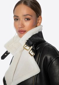 Czarna skórzana kurtka z białym kołnierzem ze shearlingu, z złotymi klamrami i zapięciem na zamek błyskawiczny. Teksturowany materiał kontrastuje między użytymi tkaninami.