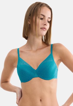 Vrouw met kort blond haar, gekleed in een teal beugelbeha met subtiel geometrisch patroon, staand tegen een effen witte achtergrond.