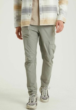Homme portant un pantalon cargo gris clair, une veste à carreaux beige et grise, et des baskets blanches et grises, debout devant un fond clair uni.