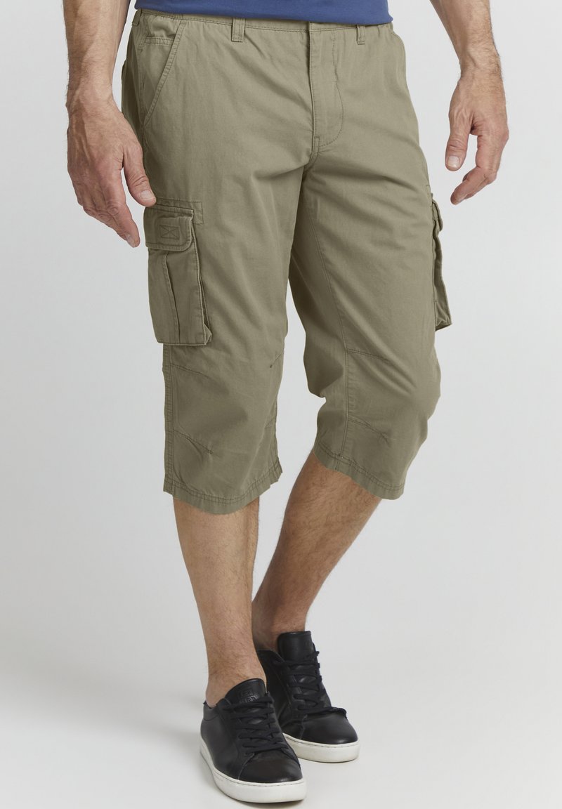 Shorts cargo en tissu vert olive clair, avec une coupe jusqu'aux genoux, dotés de poches latérales et d'une coupe décontractée, accompagnés de baskets noires.