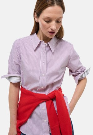 Chemise à rayures boutonnée de couleur violet clair et blanc, avec manches retroussées révélant un motif rayé contrastant. Pull rouge noué à la taille.