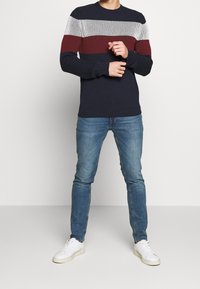 Marinblå tröja med rund hals som har horisontella ränder i grått, rött och vitt. Modellen matchar den med ljusblåjeans och vita sneakers.