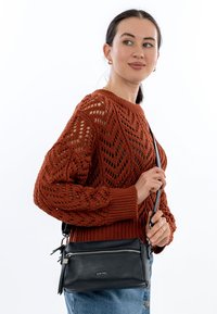 Borsa a tracolla in pelle nera con zip argentata, tracolla staccabile e superficie testurizzata; il modello indossa un maglione di lana arancione.
