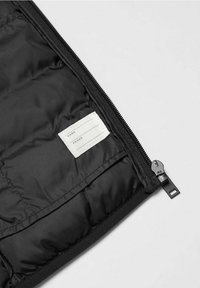 Schwarze Pufferjacke Innenansicht mit glatter Textur, steppender Polsterung und einem Stoffetikett mit Linien für Name und Telefonnummer.