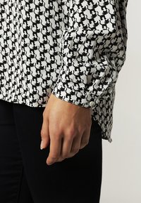 Černobílá košile s dlouhým rukávem zdobená opakovaným vzorem koček. Tkanina je hladká a má knoflíkovou aplikaci na manžetách.