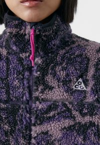 Pullover in fleece nelle tonalità di viola con un motivo astratto nero. Presenta un colletto alto, chiusura a mezza zip e un logo triangolare sul petto.