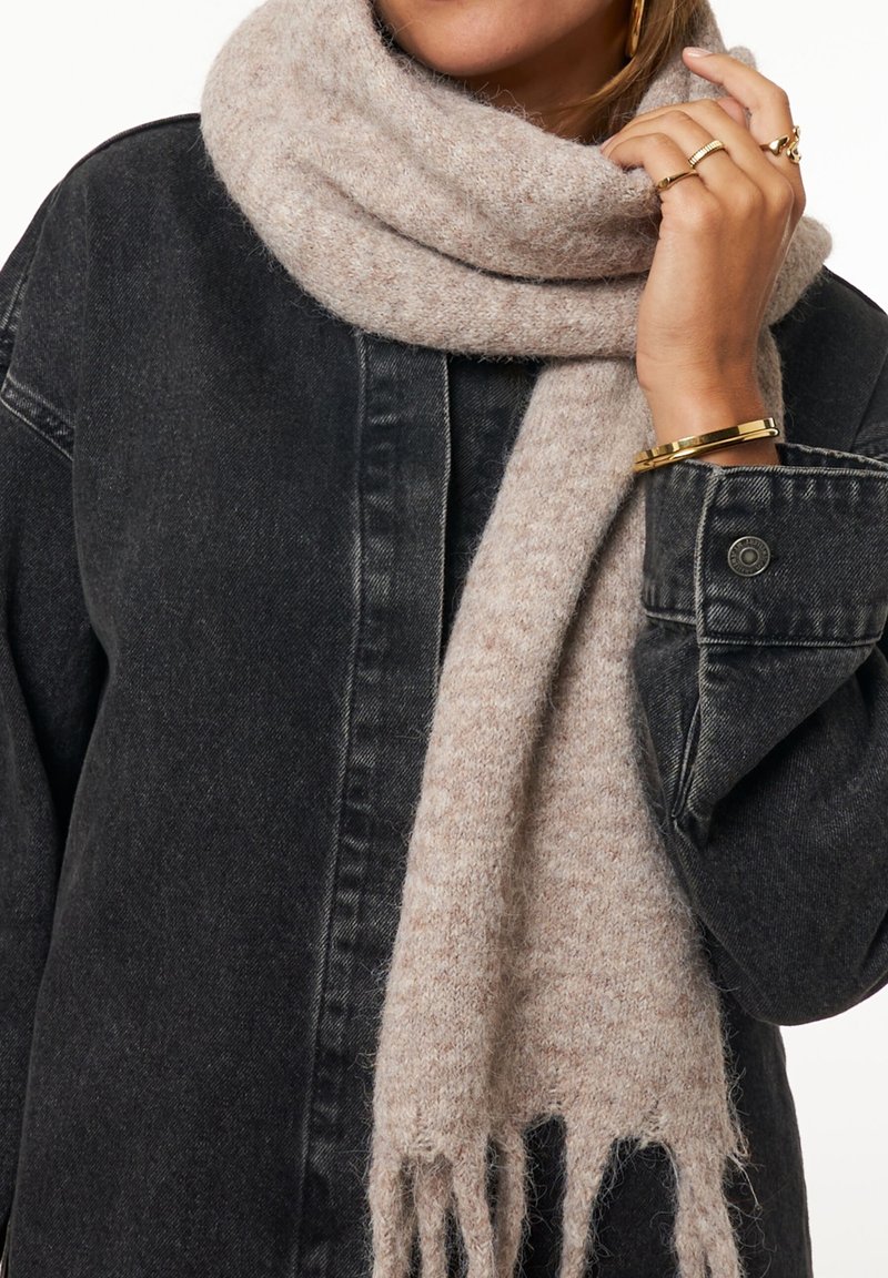 Personne portant une écharpe en tricot beige et une veste en jean noire, avec des bagues en or et un bracelet en or à la main en train d'ajuster l'écharpe.