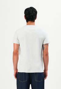 Homme aux cheveux bruns portant un t-shirt léger gris clair à manches courtes et un jean bleu foncé, se tenant de dos face à la caméra.