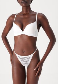 Weißer gepolsterter BH aus glattem Material mit verstellbaren Trägern, kombiniert mit einem weißen Spitzen-Tanga mit floralen Mustern und transparenten Einsätzen.