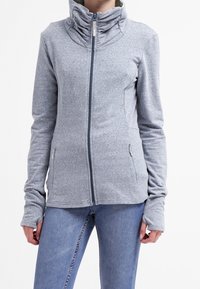 Giacca grigia con zip, colletto alto e polsini con foro per il pollice. Presenta un tessuto morbido, due tasche frontali e un design aderente.