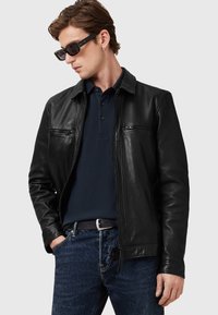 Schwarze Lederjacke mit Reißverschluss, zwei Brusttaschen und Kragen; getragen über einem marineblauen Poloshirt und Jeans.