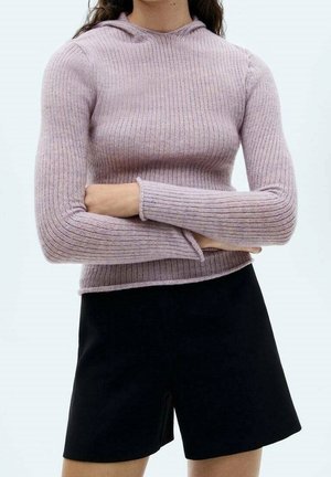 Vrouw draagt een geribbelde lichtpaarse lange mouw trui met capuchon en zwarte shorts met hoge taille, armen gekruist over de torso.