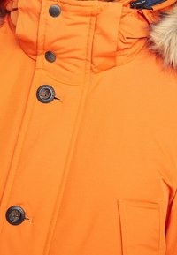 Veste d'hiver orange vif avec des boutons noirs, une capuche ornée de fausse fourrure, et une poche avant sur le côté droit.