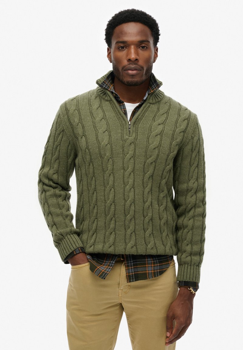 Pull en maille verte avec col à demi-zippé, porté sur une chemise à carreaux. Le pull présente des motifs texturés et des poignets côtelés.