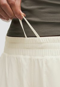 Pantalone krem boje s elastičnim pojasom i teksturiranom površinom, sa špagom za podešavanje, prikazani izbliza s rukom koja drži konopac.