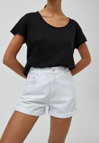 Femme portant un top noir ample à manches courtes rentré dans un short blanc taille haute à ourlets roulés, debout les mains dans le dos.