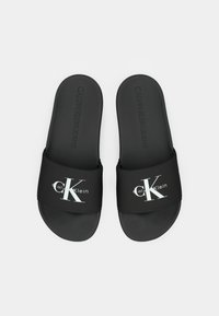 Sandales slides noires Calvin Klein avec logo blanc "CK" sur les brides, semelles intérieures rembourrées et texte embossé "Calvin Klein Jeans" à l'intérieur des semelles.