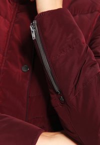 Vinröd dunjacka med dragkedjeförsedd ärmficka, knäppning med tryckknappar, quiltat mönster och ribbade muddar för extra värme.