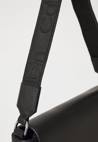 Borsa nera con superficie texturizzata, caratterizzata da una tracolla in tessuto impreziosita dal nome del marchio, accenti metallici e una forma cilindrica elegante.