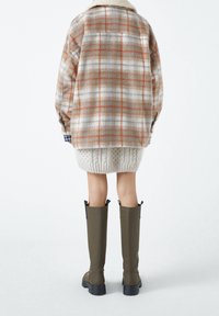 Giacca camicia a quadri beige e arancione con colletto in shearling, abbinata a un maglione a maglia cable color crema e stivali verdi fino al ginocchio.