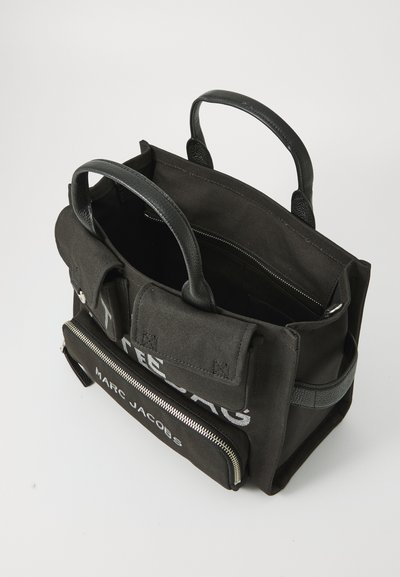 Black canvas tote bag with leather accents, top handles, front zip pocket, and printed text. Interior shows spacious compartments and lining.