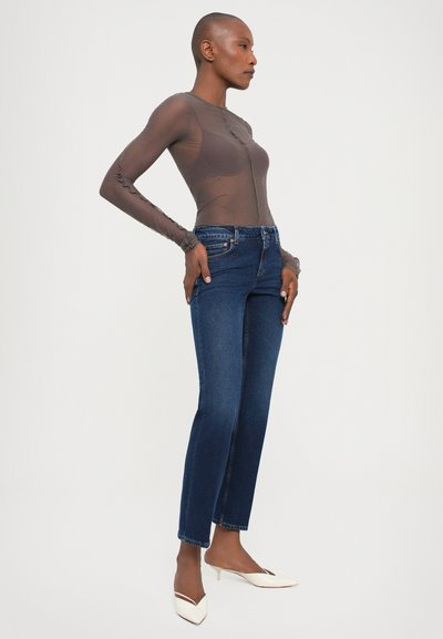 Brown sheer long-sleeve top with ruffled edges paired with dark blue straight-leg jeans. Model wears white pointed-toe heeled mules.