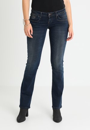 Vrouw draagt donkerblauwe slim-fit jeans met voorzakken, zwarte enkellaarsjes met hak en een zwarte top, staand tegen een effen witte achtergrond.