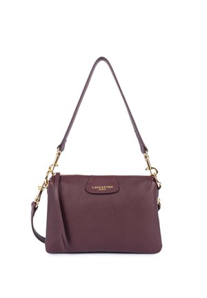 Sac à main Lancaster Paris en cuir violet foncé avec des garnitures dorées, une bandoulière et un zip avec pompon sur fond blanc.