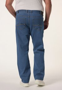 Jeans de mezclilla azul con un corte recto, que cuentan con dos bolsillos traseros y detalles de costura amarilla. Llevados con un cinturón y zapatos de color claro.