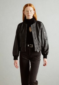 Chaqueta bomber de cuero negro con cierre de cremallera, mangas con puños y un bolsillo lateral. Lleva puesta una camiseta de cuello alto negra y se combina con jeans oscuros.
