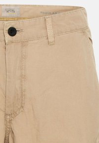 Pantalon beige décontracté avec bouton noir, passants de ceinture, coutures visibles et étiquette « Camel Active » à l’intérieur de la ceinture.