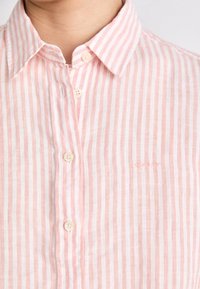 Jasnoróżowa koszula w paski, wykonana z lnu, z kołnierzykiem, perłowymi guzikami i haftowanym logo po lewej stronie.