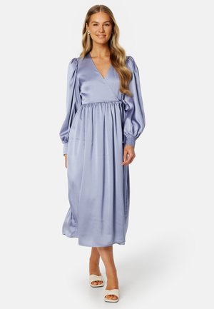 Lila satijnen wikkeljurk met pofmouwen, getailleerde taille en vloeiende rok. Heeft een V-halslijn en een enkel-lange zoom. Witte blokhak sandalen.