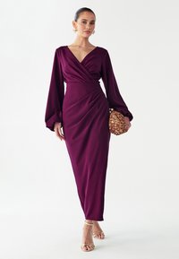 Lange paarse jurk met een diepe V-hals, overslagontwerp en volumineuze mouwen. Draagt bijpassende gouden sandalen met bandjes en houdt een gouden clutch met textuur vast.