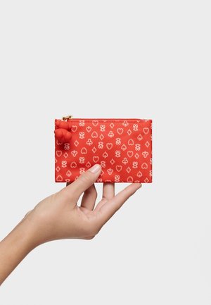 Mano sosteniendo una pequeña bolsa rectangular roja con un patrón de naipes blancos, con un tirador de cremallera en forma de oso rojo.