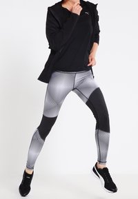 Sort træningsjakke over en tætsiddende sort top og leggings med sort-hvide mønstre, der har geometriske designs og teksturerede detaljer. Sorte sneakers.