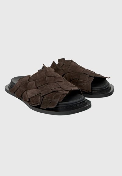 Tmavě hnědé semišové sandály s pletenými pásky a černými plochými podrážkami, navržené pro snadné nazouvání s otevřenou špičkou.