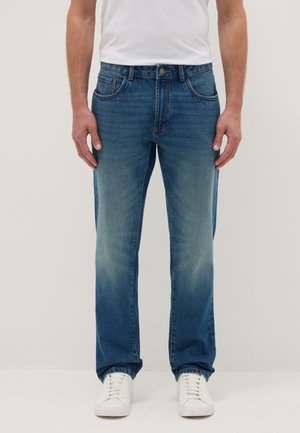 Mann trägt gerade geschnittene blaue Jeans mit verblasster Vorderseite und weißen Sneakers, steht vor einem schlichten hellen Hintergrund.