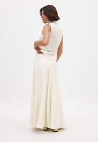 Lång, flödande klänning i off-white med en texturerad, smockad bodice och hög krage. Kjolen faller mjukt ner till golvet. Enkelt, elegant design.