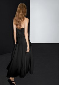 Zwarte strapless-maxi-jurk met een vloeiende rok, een gladde textuur en een getailleerde top, gedragen met platte sandalen tegen een donkere achtergrond.