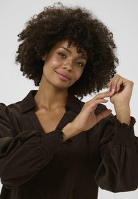 Blouse en velours côtelé marron avec un décolleté en V, des manches bouffantes avec des accents smockés aux poignets, et une texture lisse.