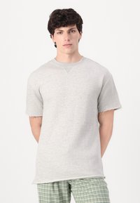 Pulover gri cu mânecă scurtă, cu tivuri și margini nefinisate, având un decolteu tip crew și o textură moale, purtat peste pantaloni în carouri.