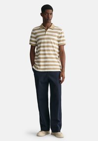 Polo de rayas en beige y blanco, de mangas cortas, con cuello, combinado con pantalones oscuros en marino y zapatos claros. Fondo uniforme.
